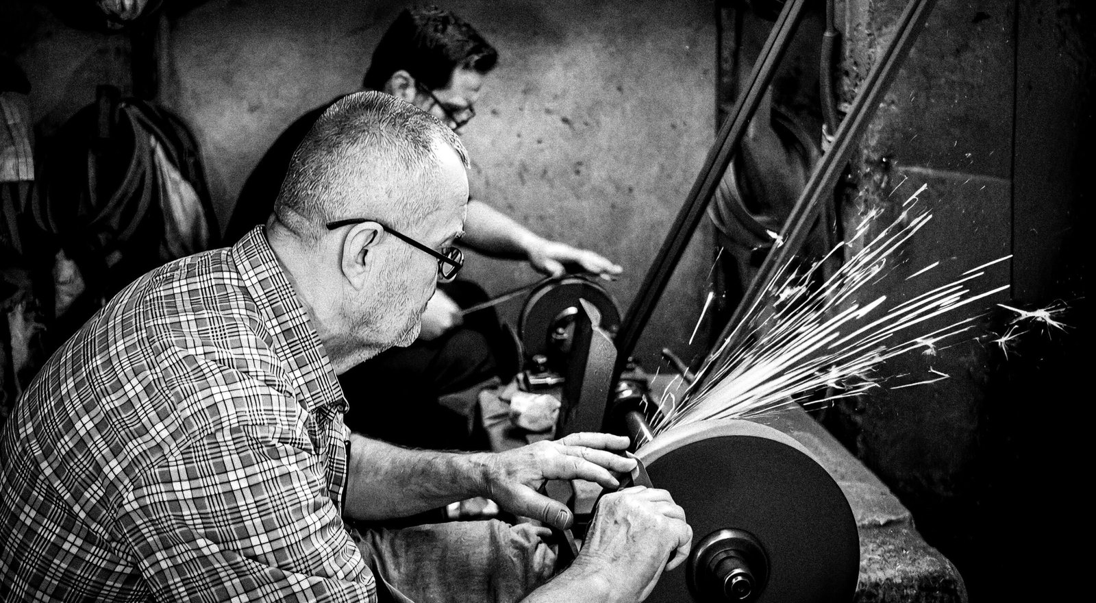 Two craftsmen sharpening tools on grinders, sparks flying, in a workshop.