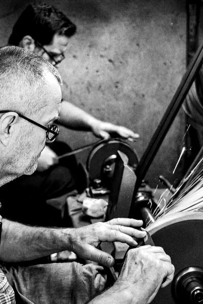 Two craftsmen sharpening tools on grinders, sparks flying, in a workshop.